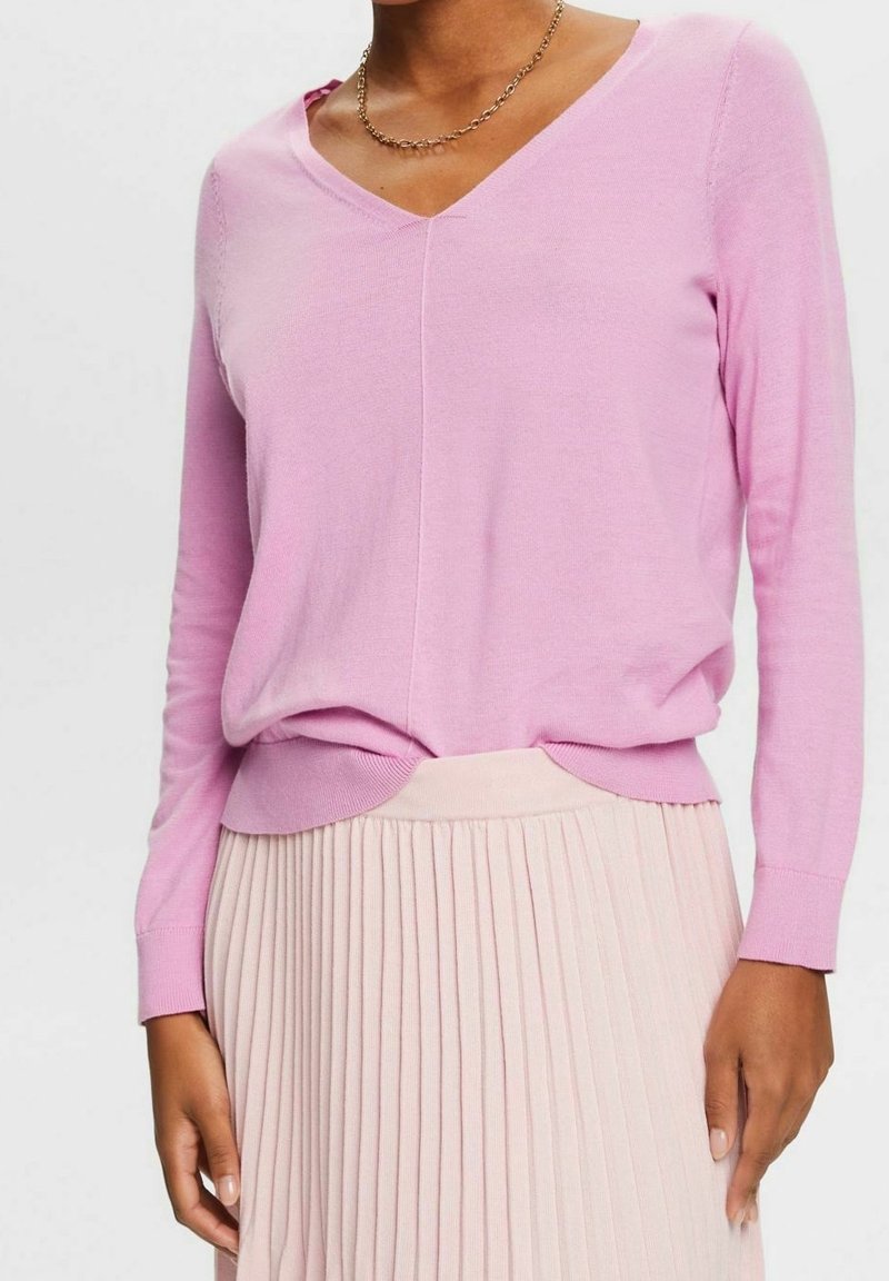 Femme portant un pull à col en V rose clair et une jupe plissée rose pâle, avec un collier chaîne en or visible.