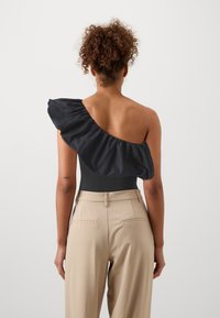 Melna bodysuit ar vienu plecu un viļņotu piedurkni, apvienots ar augsto vidukli bežām biksēm, kurām ir aizmugurējās kabatas un gluda tekstūra.