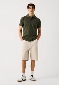 Olivengrøn polo skjorte med korte ærmer og krave; beige shorts, knælange; hvide sneakers med sorte detaljer; modellen står med hænderne i lommerne.
