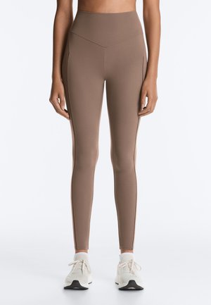 Femme portant un legging marron taille haute et des baskets blanches, debout les bras détendus le long du corps sur fond blanc.