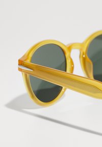 Las gafas de sol redondas tienen un marco transparente amarillo, lentes verdes oscuros y varillas amarillas a rayas con acentos plateados.