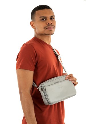 Sac bandoulière gris avec un motif texturé, bandoulière ajustable et compartiments zippés, porté par une personne vêtue d'une chemise couleur rouille.