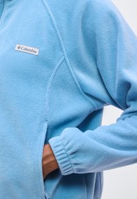 Χέρι στην τσέπη ενός γαλάζιου fleece μπουφάν Columbia με ελαστική μανσέτα και ορατό λογότυπο στη θώρακα.