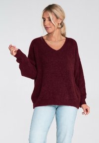 Kobieta z blond włosami w kucyku, nosząca złote kolczyki w kształcie kół, złoty naszyjnik, bordowy sweter z dekoltem w serek oraz jasnoniebieskie dżinsy.