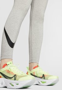 Šedé legíny s černým logem Nike, doplněné neonově žlutozelenými a oranžovými teniskami s masivní bílou podrážkou a černými akcenty.