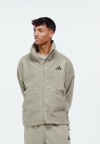 Man wearing light beige zip-up fleece jacket with high collar and matching pants, looking sideways against plain white background.