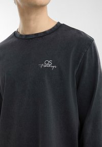 Dunkelgraues Langarmshirt aus weichem Stoff mit Rundhalsausschnitt und kleinem weißen gestickten Logo auf der Brust.