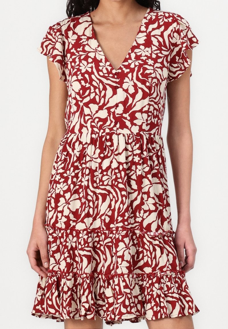 Vestido floral rojo y crema con escote en V, mangas cortas con volantes, costura en la cintura y un dobladillo con volantes en capas. Tejido ligero con un acabado texturizado.