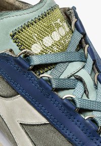 Zapatilla de múltiples materiales con un detalle tejido en verde, acentos en azul claro, tela gris y cuero azul marino. Incluye ojales de metal para los cordones.