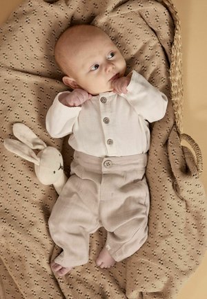 Bambino sdraiato su una coperta beige lavorata a maglia all'interno di un cesto intrecciato, indossando un completo color crema, che guarda di lato, con un morbido peluche a forma di coniglio nelle vicinanze.