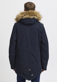 Marine parka jakke med hætte i pels, der har lynlåslukning foran, sidelommer og en længere rygkant med en trekantet detalje.
