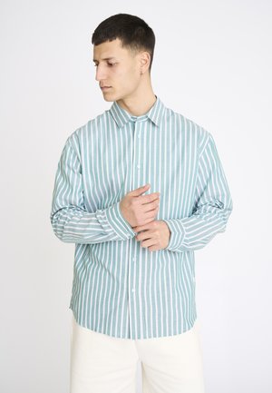 Ung mand med kort mørkt hår iført en teal og hvid vertikalt stribet skjorte med knapper og lyse bukser, der justerer sin ærme.