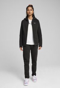 Sudadera negra con cremallera y capucha, combinada con joggers negros ajustados. Una camiseta blanca por debajo, complementada con zapatillas coloridas.