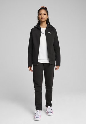 Sudadera negra con cremallera y capucha, combinada con joggers negros ajustados. Una camiseta blanca por debajo, complementada con zapatillas coloridas.