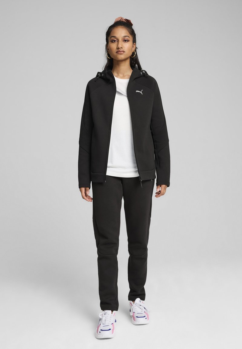 Sudadera negra con cremallera y capucha, combinada con joggers negros ajustados. Una camiseta blanca por debajo, complementada con zapatillas coloridas.