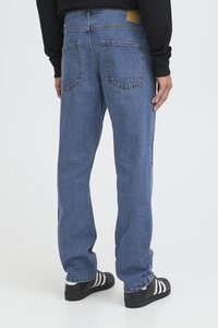 Persoană îmbrăcată cu blugi drepti albaștri și adidași negri cu dungi albe, stând pe un fundal deschis simplu.