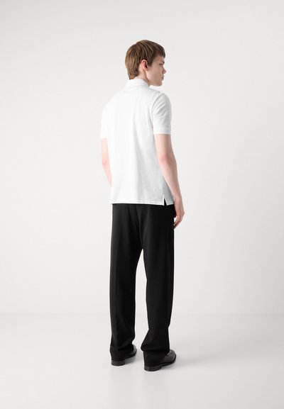 Polo blanc à manches courtes, avec des fentes latérales, assorti à un pantalon noir ample et des chaussures noires, vu de dos.