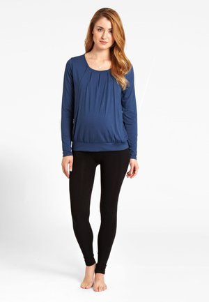 Námornícky modré tričko s dlhým rukávom a skladaným designom, doplnené čiernymi legínami. Mäkký materiál s hladkou textúrou, priliehavý strih.