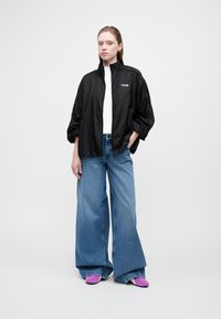 Ung kvinde iført en sort zip-up jakke, hvid bluse, brede blå jeans og lilla sko, stående mod en ensfarvet lys baggrund.