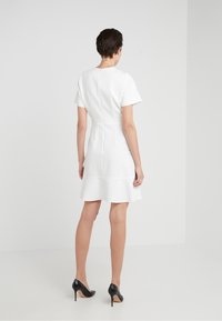 Femme aux cheveux courts et foncés, portant une robe blanche à manches courtes et des talons noirs, debout, dos tourné, sur un fond blanc uni.