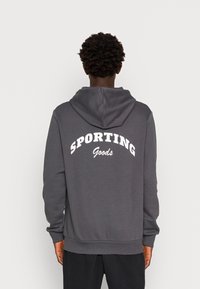 Felpa grigia con cappuccio regolabile, polsini e orlo a costine. Sul retro presenta un grande testo bianco che recita "SPORTING Goods" in un design arcuato.