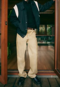 Czarna kurtka z białymi akcentami, czarna koszula, beżowe luźne spodnie i czarne mokasyny. Drewniana podłoga i zieleń w tle.