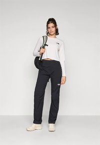 Νεαρή γυναίκα που στέκεται φορώντας ένα λευκό crop top με μακριά μανίκια, μαύρο παντελόνι, κρεμ αθλητικά παπούτσια και ένα μαύρο σακίδιο στον έναν ώμο.