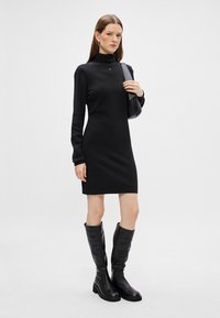 Vestido preto justo com gola alta, mangas longas e um logótipo subtil. Usado com botas altas de couro preto. Textura suave, design minimalista.