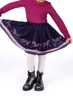 Dítě v tmavě modré sukni s motivem koní, bílých punčochách, černých botách a dlouhém rukávu vínového topu, držící sukni široce rozevřenou.