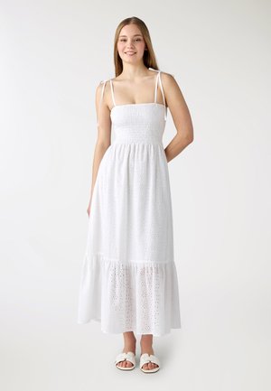 Giovane donna sorridente, che indossa un abito midi bianco senza maniche con occhielli e lacci sulle spalle, e sandali bianchi a ciabatta, in piedi davanti a uno sfondo semplice.