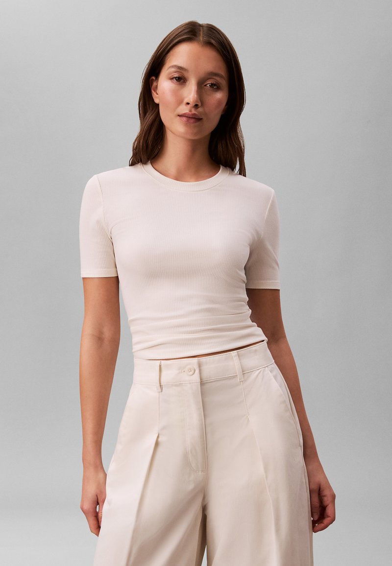 Femme aux longs cheveux bruns portant un haut blanc ajusté à manches courtes et un pantalon large crème taille haute sur un fond gris uni.