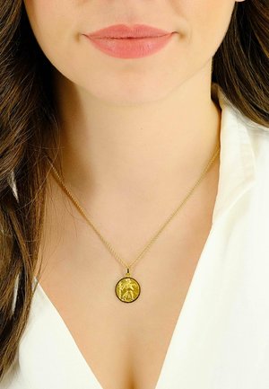 Collana con pendente d'oro, caratterizzata da un charm circolare con un design a rilievo dettagliato, sospesa su una fine catena, indossata sopra una blusa bianca.
