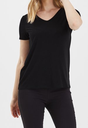 Kobieta w prostym czarnym T-shircie z krótkim rękawem i dekoltem w serek oraz czarnych spodniach na jednolitym białym tle.