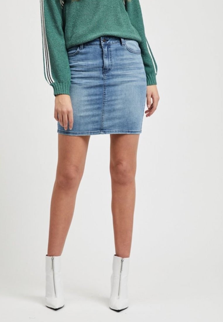 Jupe mini en denim bleu clair avec une coupe classique et un dégradé subtil, associée à un pull vert. Elle comprend une poche avant et une fermeture éclair.