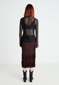 Femeie cu păr roșu până la umeri, purtând un top lung cu mâneci lungi din material transparent de culoare neagră, sutien negru, fustă midi plisată de culoare maro și ghete negre cu gleznă, stând.
