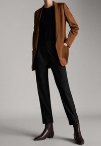 Braunes Sakko über einem schwarzen Oberteil, kombiniert mit schwarzen Hosen und braunen Stiefeletten. Glatte Textur, maßgeschneiderte Passform und aufgekrempelte Säume an den Hosen.