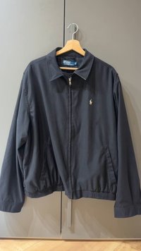 Black jacket with a zip front, soft fabric, spread collar, and elastic waistband. Features a small logo on the chest. Hangs on a wooden hanger.