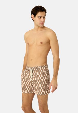 Skjorteløs mand iført brune badeshorts med hvidt geometrisk mønster og snøre, stående mod en simpel lys baggrund og kiggende til siden.