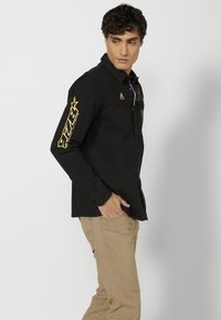 Camisa negra de botones hecha de algodón, con acentos dorados y gráficos en las mangas, presenta un corte ajustado y un cuello puntiagudo.