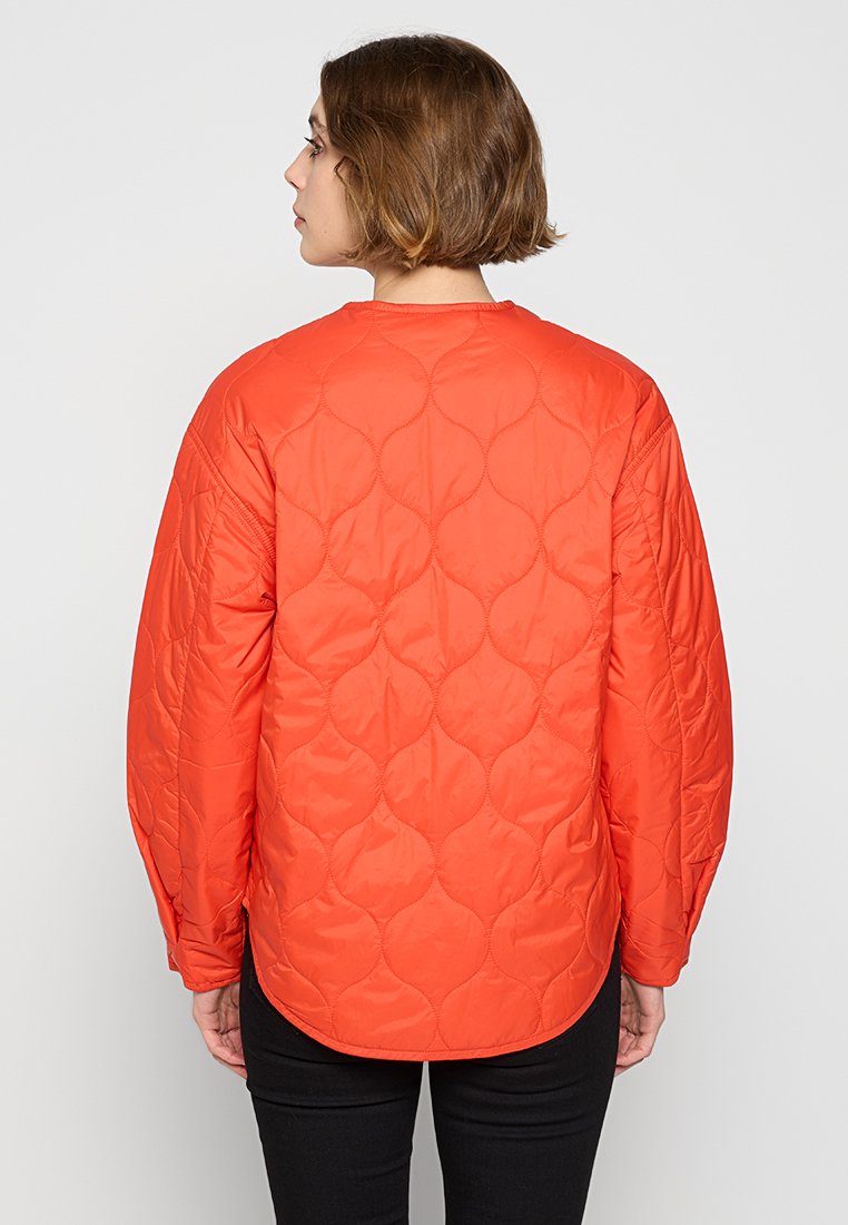 Pomarańczowa pikowana kurtka z okrągłym dekoltem i długimi rękawami, z teksturowanym wzorem na tkaninie. Widok z tyłu z lekko zakrzywionym dołem.