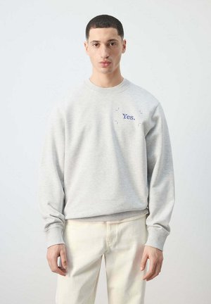 Joven con cabello corto y oscuro que lleva una sudadera gris claro con "Yes." impreso en azul en el pecho y pantalones color hueso.