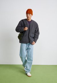 Giacca in pile grigia con zip frontale, due grandi tasche frontali, abbinata a jeans azzurri e un berretto color ruggine, oltre a una borsa a tracolla verde.