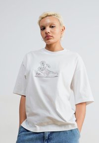 T-shirt en coton blanc avec un graphisme de sneaker agrémenté d'éléments floraux. Coupe décontractée avec des manches courtes et un col rond.