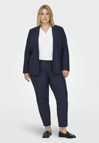 Námořnický modrý blazer a kalhoty na míru vyrobené z elastické látky, kombinované s bílým topem. Blazer má klopový límec a dvě přední kapsy.