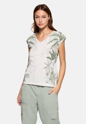 Mujer con una blusa blanca de cuello en V con estampados de hojas tropicales verdes y pantalones cargo verde claro, de pie con una mano en el bolsillo.