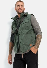 Grüne Weste mit mehreren Taschen, reißverschluss vorne und Kapuze. Getragen über einem grauen Langarmshirt. Verfügt über Stichdetails und ein Lederetikett.