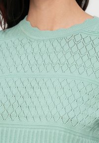 Ανοιχτό πράσινο πλεκτό πουλόβερ με υφασμένο διαμάντι μοτίβο και ριμπ τελείωμα. Ο λαιμός είναι κυματιστός. Λείο ύφασμα με ελαφριά διαφάνεια.