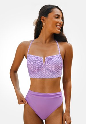 Femme souriante, portant un haut de bikini style licou à fleurs violettes et un bas uni taille haute, avec de longs cheveux et des boucles d'oreilles en forme d'étoile.