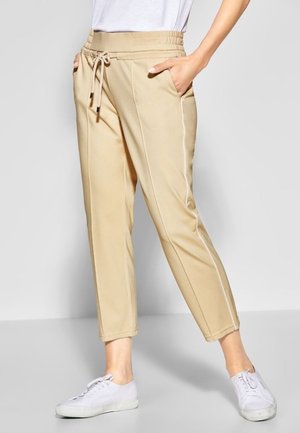 Persoon draagt beige broek met trekkoord en zijnaden en witte sneakers, staand met handen in de zakken tegen een grijze achtergrond.