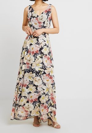Długa sukienka na ramiączkach z dekoltem w serek, z dużym kwiatowym wzorem w kolorach kremowym, różowym i żółtym na czarnym tle, noszona z złotymi sandałami na wysokim obcasie.
